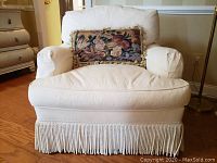 Front view of cream textured upholstered chair with floral throw pillow and twisted fringe trim at the base, showing some stains.