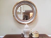 Front view of round mirror hanging on wall with lidded urn, two candles in holders, and decorative bowl on table below.