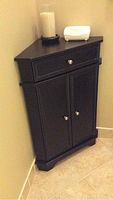 Black corner cabinet from front with drawer and storage closed, positioned in a corner on tiled floor.