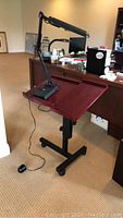 Adjustable wooden podium with dark reddish-brown finish and black base with wheels, with a black adjustable desk lamp placed on top, shown in an office setting.