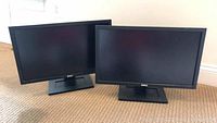 Two Dell monitors side by side, black frame and rectangular base stands shown on carpet.