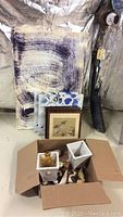 Photo showing stacked framed and unframed paintings and prints with shore bird figurines and ceramics packed in a box on the floor.