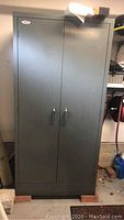 Front view of tall gray metal cabinet with double doors and handles, standing on wooden blocks.