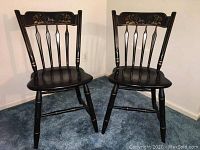 Two black painted wooden thumb back chairs with deer stenciled top rail and gold accent paint on legs and backrest edges, placed on blue carpet.