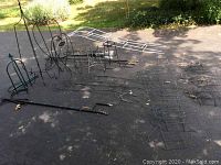 Wide view of assorted metal garden items including plant stands, shepherd's hooks, and trellises laid out on pavement with some shade.