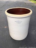 Stoneware crock standing on asphalt showing its white exterior with a blue crown and number 12 on the front.