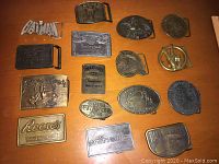 Top-down view of 15 advertising belt buckles laid out on a wooden surface showcasing different brands and designs.