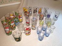 Full view of 28 collectible drinking glasses with various cartoon characters from Camp Snoopy, Looney Tunes and others laid out on a flat surface.