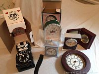 Photo showing the full assortment of clocks including Kit-Cat clock, kitchen clock in box, two Howard Miller desktop clocks, miniature mantle clock, and round wall clock.