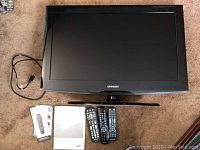 Samsung 32-inch flat panel television on carpet with four remotes and user manual positioned in front.