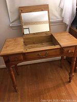 Wooden makeup table with center lift-up top revealing mirror and storage, side drawers visible, standing on hardwood floor.