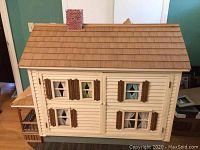 Exterior front view of doll house showing cream painted walls, wood-shingled roof, brick chimney and shuttered windows