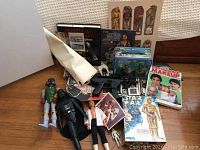 Wide photo showing a collection of Star Wars figures including Darth Vader, Luke Skywalker, Boba Fett, toys, comics, games and print materials on a wooden floor.