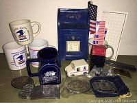 Overview of USPS memorabilia items including mugs, collectible USPS badges, small USPS letter collection box replica, and USPS mail truck model on a wooden surface.