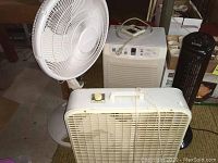 Photo showing all four items: white pedestal fan, white box fan in front, white dehumidifier at the back, and black tower fan to the right.