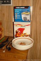 Black & Decker rice cooker box on top with tomato press box below, four stacked ceramic pasta bowls with bird motif in front, assorted knives with wooden handles laid out beside them.