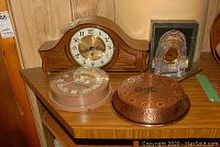 Photo showing four clocks on a wooden surface including a wood mantle clock, copper fry pan clock, Westclox plastic clock, and glass desk clock