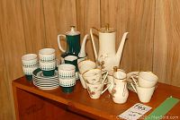 Two espresso sets side by side on wooden shelf showing cups, saucers, pots, creamer, and sugar bowl.