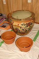 Large Asian motif ceramic planter with intricate designs of cranes, trees, and mountains shown together with two small terra cotta pots on a cloth.