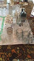 Photo showing assorted clear glassware including beer steins, shot glasses, and some tall glasses, alongside Royal Albert floral china tea cups and saucers and a black train bottle holder visible in background.
