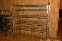Front view of chrome metal shelving unit with five wire shelves on wheels in wood-paneled room.