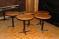 Five round restaurant style bistro tables with woodgrain melamine tops and black metal pedestal bases arranged in a room with wood flooring and brick wall.