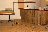 Full view of the rolling metal clothing rack in a room, showing the rectangular metal frame with a horizontal hanging bar and four caster wheels on the bottom.