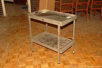Full view of vintage metal trolley with glass top shelf, lower metal shelf with decorative cutout, and bottle/glass holder on side. Caster wheels visible for mobility.