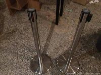 Two stainless steel stanchion posts standing on a gravel floor, showing weighted round bases and retractable belt mechanisms at the top.