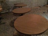 Four round restaurant style bistro tables arranged in a row on gravel surface inside a basement or storage space. Tables have brown wood grain melamine tops and black metal bases.