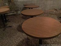Four matching round restaurant style bistro tables arranged in a row on a gravel surface. Woodgrain melamine tops and black metal bases visible.