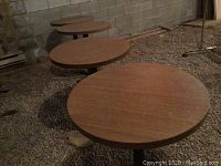 Three round wooden-look bistro tables with black metal pedestal bases on gravel floor