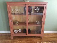 Front view of the painted wooden IKEA cabinet with two glass sliding doors displaying glassware and decorative items on shelves inside.