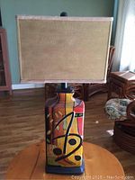 Full front view of the colorful abstract design lamp with rectangular beige shade on a round wooden table.