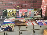 Photo of a collection of vinyl records, mostly country and classic albums, displayed on furniture in front of a brick wall.
