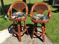 Two cane bar stools with decorative floral padded cushions shown outdoors on concrete and grass. Different heights visible.