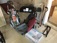 Photo showing entire lot arranged on floor near sofa and stairwell, items include shopping trolley with vegetable print, padded back support chair, cane, portable folding table, and boxed raised toilet seat.