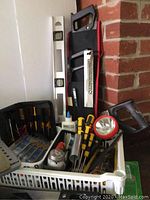 Photo of assorted hand tools including spirit level, saw, flashlight, screwdrivers, and adhesive glue bottle inside a plastic bin