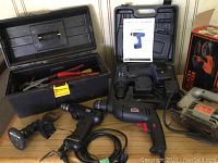 Black plastic toolbox opened showing various hand tools including pliers, wrenches, screwdrivers. Beside it are three power tools: a compact Black & Decker drill, a Black & Decker Mouse detail sander, and a corded drill.