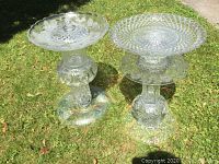Photo showing two handmade decorative glass crystal bird baths standing on grass outdoors, one taller with a raised central finial detail, both with intricate engraved patterns