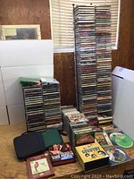 Photo showing tall stacks of assorted music CDs in jewel cases, some boxed collections, and two CD carrying cases in foreground.
