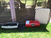 Full barbecue lot showing the Thermos Grill2Go red portable gas grill, black grill stand, and white Coleman folding table on grass outdoors under a tent.