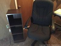 Photo showing black office chair next to small dark brown bookcase with three shelves.