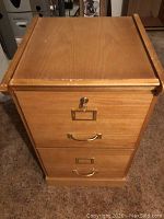 Front view of the wooden filing cabinet showing two drawers with brass handles and label holders, a lock on the top drawer with key inserted.