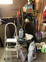 Photo of white metal step ladder, bags of crushed rock and pea gravel, various garden tools behind including shovels and rakes, tomato planter box and coiled garden hose.