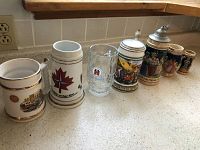 Seven beer steins and mugs lined up on kitchen countertop including German and English styles, one glass mug, and a Canadian maple leaf mug.