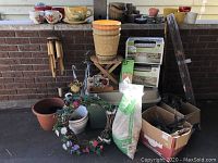 A variety of gardening items set on a porch floor include pots, wind chimes, garden border stones, a bag of soil or fertilizer, and other accessories.