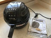 Kuraidori black air cooker with attached power cord, digital touchscreen controls on top, and recipe book beside it.