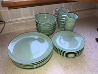Photo showing stacks of 6 dinner plates, 6 side plates, 6 bowls, and 6 mugs on a countertop in soft light green color.
