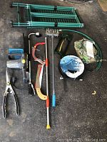 Photo showing assembled garden sprinkler, hand tools including pruning shears, cultivator, small saw, and rolled green garden hose.